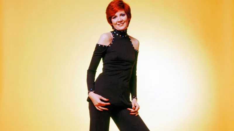 CB Prime LTD Cilla Black poses in front of a yellow background. She wears a black jumpsuit with cut out shoulders and a high neck. She has short red hair. The image is likely from the 70s era.