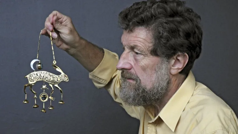 Side profile of Kit Williams, who has a beard, and is dangling the golden hare in front of his face and looking at it intently.