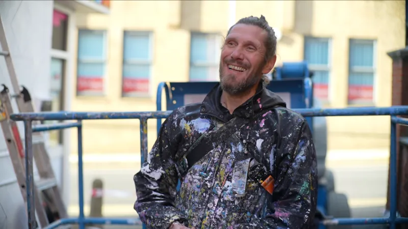 Jamie Niblock/INLIBER Artist Atma laughs as he looks away from the camera. He stands in front of a cherry picker that rests near the bottom of the building. He wears a black coat that is splattered with different coloured paint. He has long brown hair that has been tied up in a bun on top of his head and he has a thick dark beard.
