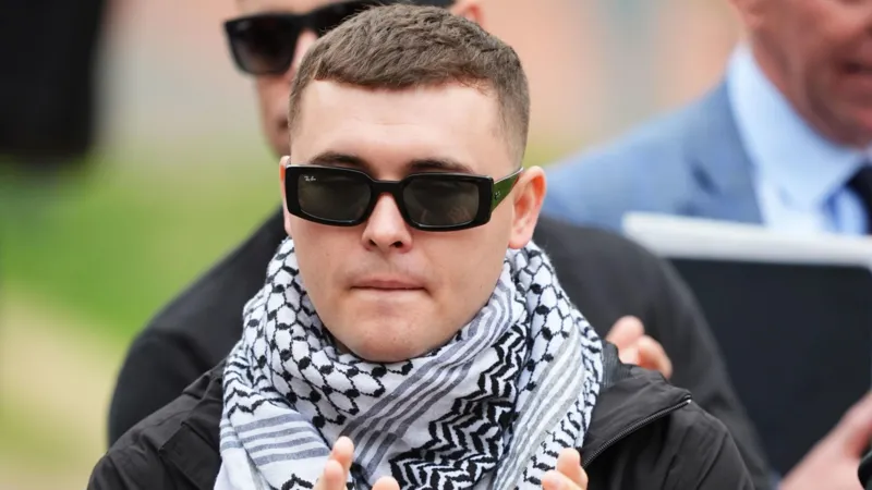 PA Media Kneecap's Liam Óg Ó hAnnaidh, who performs under the stage name Mo Chara, outside Woolwich Crown Court, London. He is wearing black sunglasses and a white and black keffiyeh. He has dark hair.