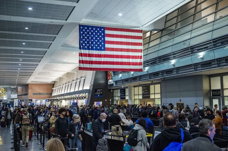 Anadolu via Getty Images Long lines of people with suitcases wrap around an airport terminal. An American flag hangs overhead.