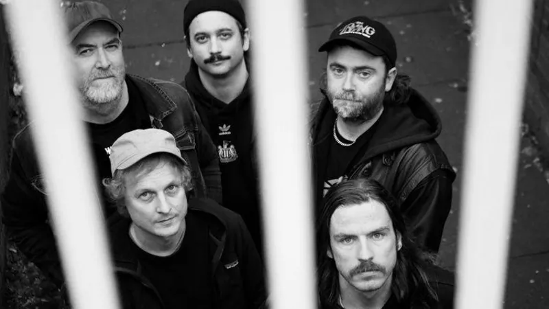 David Hall An aerial photograph of the band grouped together looking above them into the camera. There are bars in front of them. They are standing on a path and are all wearing black. The man standing in the middle is wearing a jumper with the Newcastle United club crest in the centre.