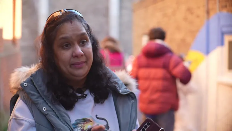 Jamie Niblock/INLIBER Anjali Sule stands at the bottom of the building where the mural is painted by artists behind her. She has long curly black hair and has sunglasses on top of her head. She wears a blue gilet jacket and a purple jumper underneath. 