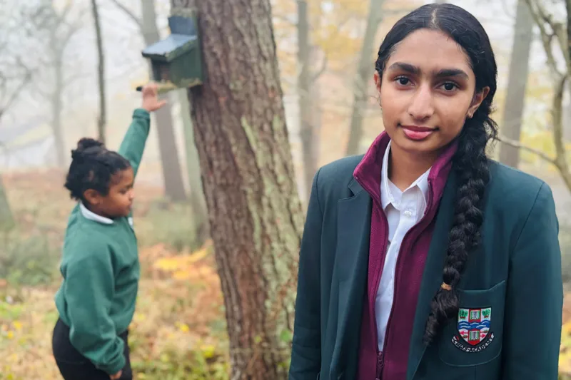 Schoolgirl sisters Avni and Vani are pictured in woodland, Vani looking at the camera, and Avni raising a hand toward a squirrel box on a tree.