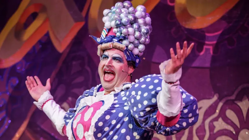 The Grove Theatre Dunstable Will Kenning, dressed as a pantomime dame with heavy makeup, wig, curlers and a bright costume.
