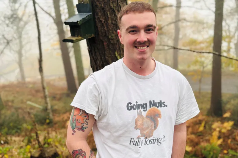 Joel Sangster wearing a 'going nuts' T-shirt featuring a squirrel, smiling at the camera, in woods; he has a squirrel tattoo on his arm.