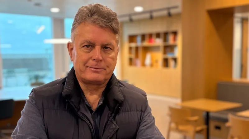 JP Allard di kantor modern, mengenakan gilet gelap, tersenyum di depan rak buku.