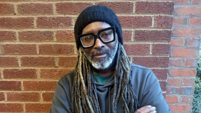 Lindsay Dube. Ia memiliki rambut panjang hitam, mengenakan jumper abu-abu tua dan topi beanie hitam, latar tembok bata berwarna merah.