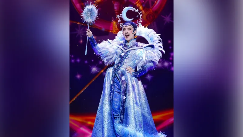 A performer on stage wearing a purple-blue dress with feathered shoulders and a moon-shaped hat