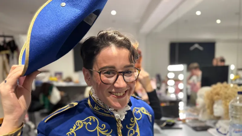 A woman with short brown hair wearing a blue costume with gold patterns. She has glasses and is tipping her hat to the camera.