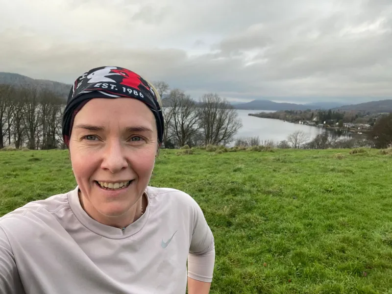 A woman with a patterned covering on her hair takes a selfie in front of a grassy field and a lake.