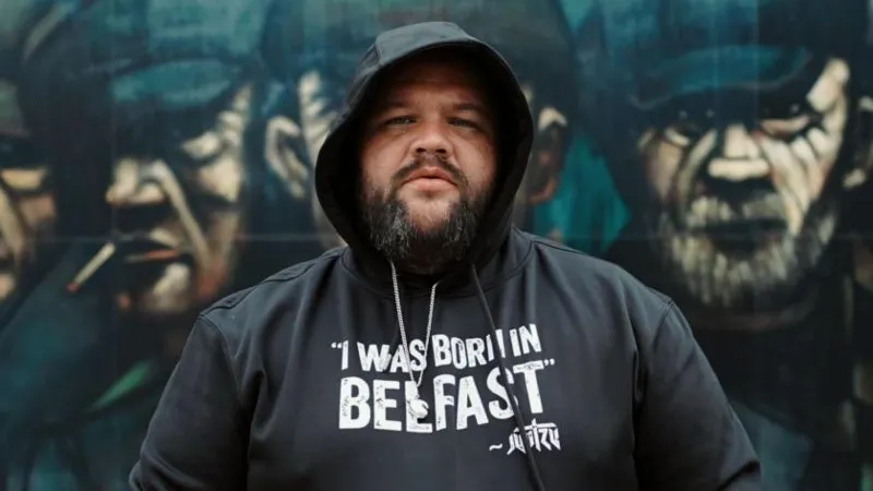 Skeet/Peter Whelan Jun Tzu. Dia memiliki janggut gelap, mengenakan hoodie hitam, dengan tulisan 'I Was Born In Belfast' di bagian depannya. Dinding mural berisi pria-pria memakai topi news