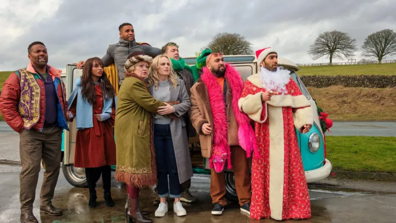A group of people standing together in front of a turquoise and white camper van on a wet road, wearing winter clothes