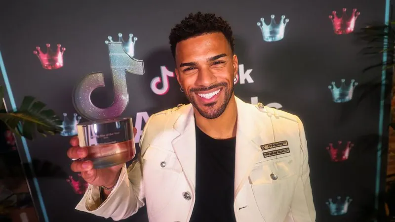Getty Images A man in a white embellished jacket smiles at the camera, holding a sparkly glass award in the shape of the TikTok logo.
