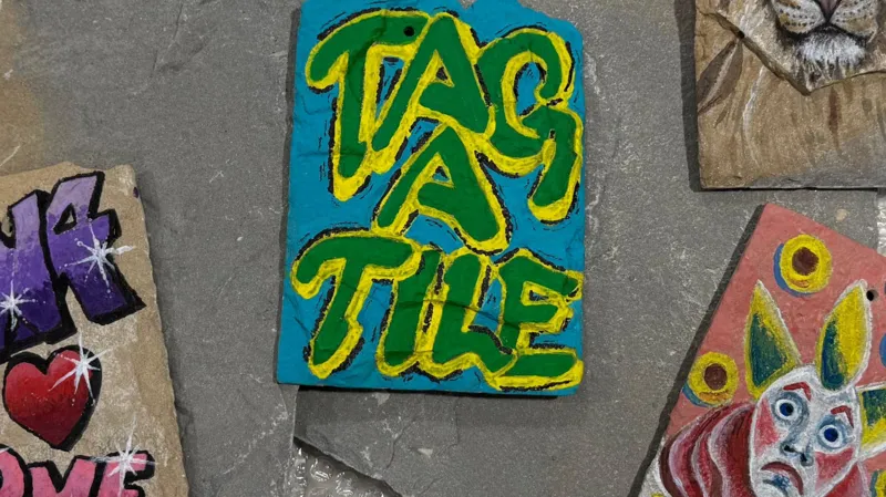 Cornerstone Four tiles on a floor, painting in different styles and colours. One, in the middle, says Tag A Tile, in green and yellow writing on a blue background. One has a sad clown's face on it, another to the left, with writing on and one at the top right, a lion. 