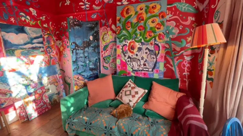 Qizil devorli xona, divanda qoramtir yashil rangli divan, yonida mushuk; devorlar gullar bilan bezatilgan.