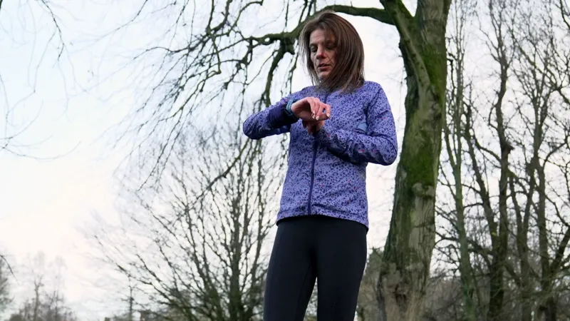 Kristian Johnson/INLIBER A woman with brown hair in a purple sports jacket stands in a park, looking at her smartwatch.