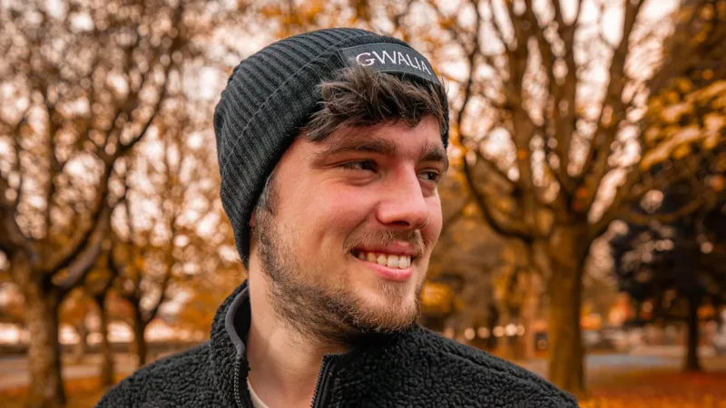 Cai Rhys Photograph of Cai Rhys, 26, taken in a autumnal park. Faded bare trees can be seen in the background, with orange leaves covering the floor. He wears a black beanie hat reading