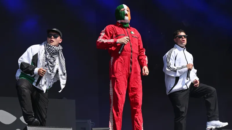 Mo Chara, DJ Próvaí, dan Móglaí Bap di panggung Glastonbury pada bulan Juni. Mo Chara dan Móglaí Bap mengenakan jaket olahraga putih, sedangkan DJ Próvaí berdiri di antara mereka dengan balaclava merah, hijau, putih, dan oranye.