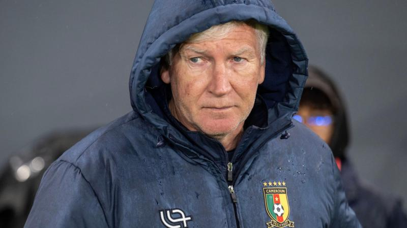 Marc Brys is seen from chest up wearing a dark blue rain jacket, with a Cameroon crest on the left breast, with the hood up. Wet patches on the jacket indicate the rain that is falling