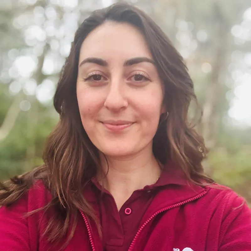 Naomi Bentley of Saving Scotland's Red Squirrels is photographed in a bright maroon zip top with the wording 'Saving Scotland's Red Squirrels' on it, smiling at camera, in woods.