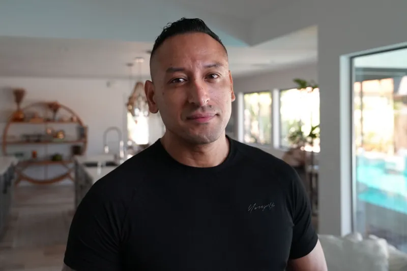 Tony Marquez stands in what appears to be a living room of a home. He is wearing a black T-shirt