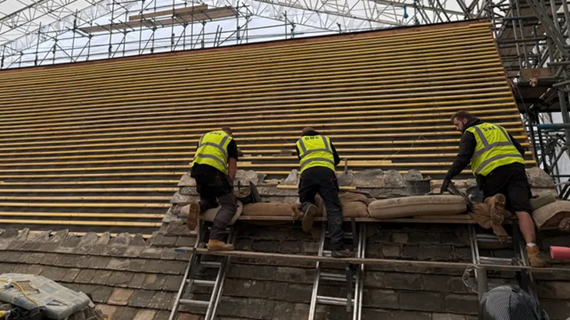 North Northamptonshire Council Three men, in high-vis jackets, working on a roof. There are three ladders, a roof with lots of wooden slates on it and scaffolding across the whole top. Slates are below the men. 